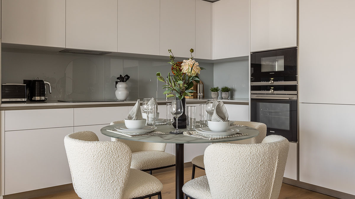 A modern minimalist kitchen and dining area by InStyle Direct featuring white handleless cabinetry, integrated Siemens appliances, and a marble-top dining table with white bouclé chairs and elegant place settings 