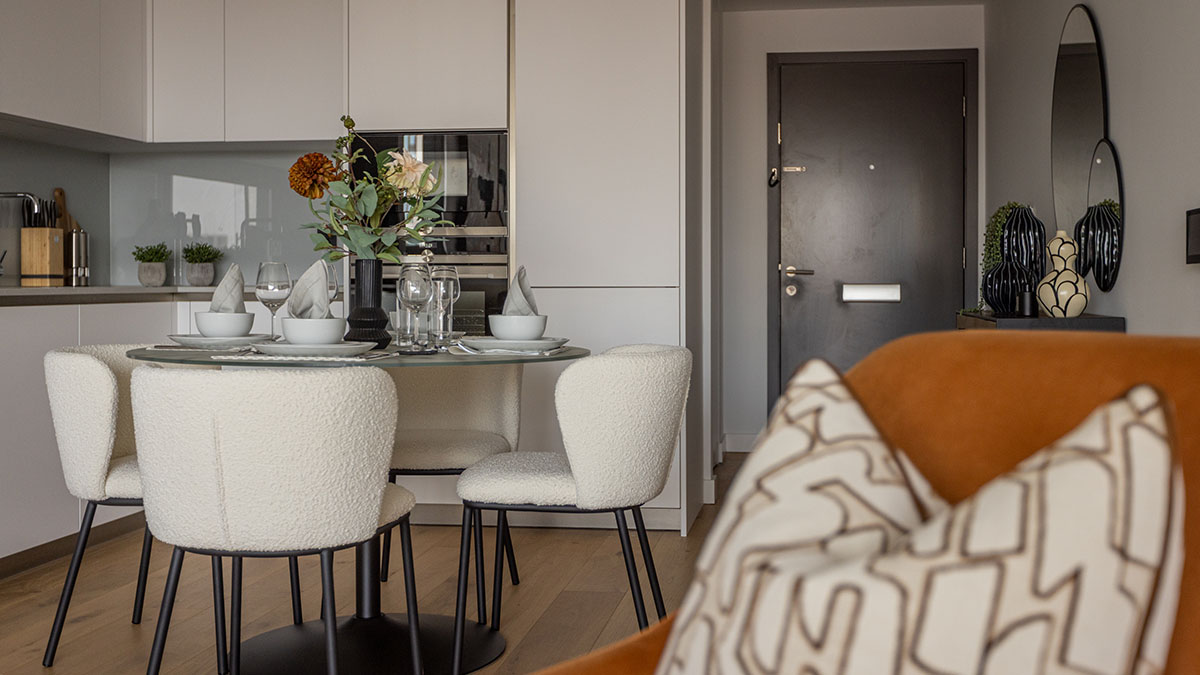 An open-plan apartment view showing a dining area with bouclé chairs leading toward a dark wood entrance door, featuring bespoke decor like a black ribbed vase and a circular wall mirror styled by InStyle Direct