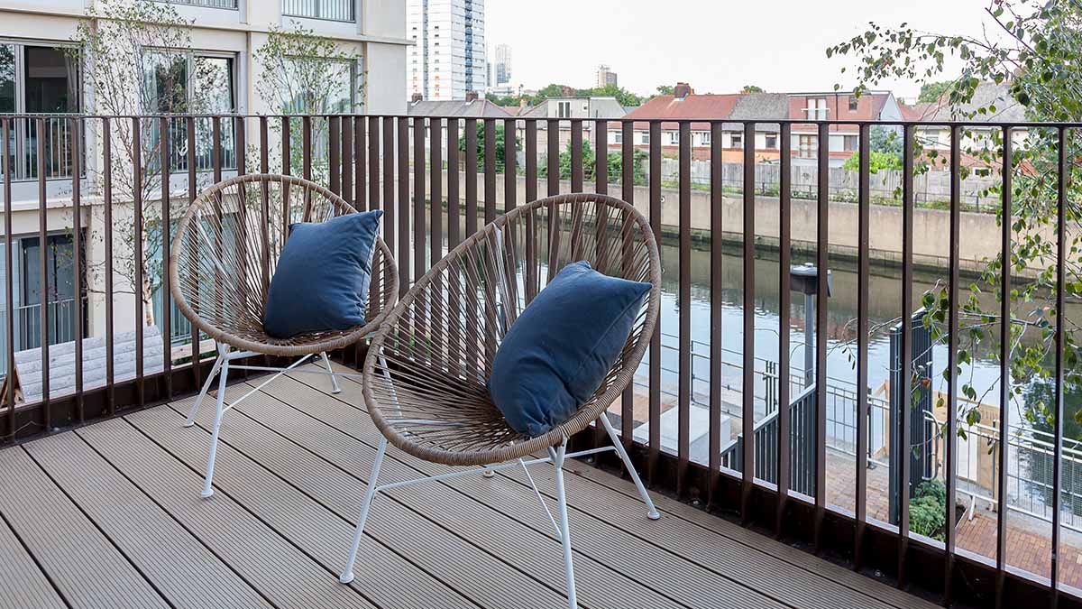 Apartment balcony with two rattan chairs and cushions overlooking a canal and residential buildings