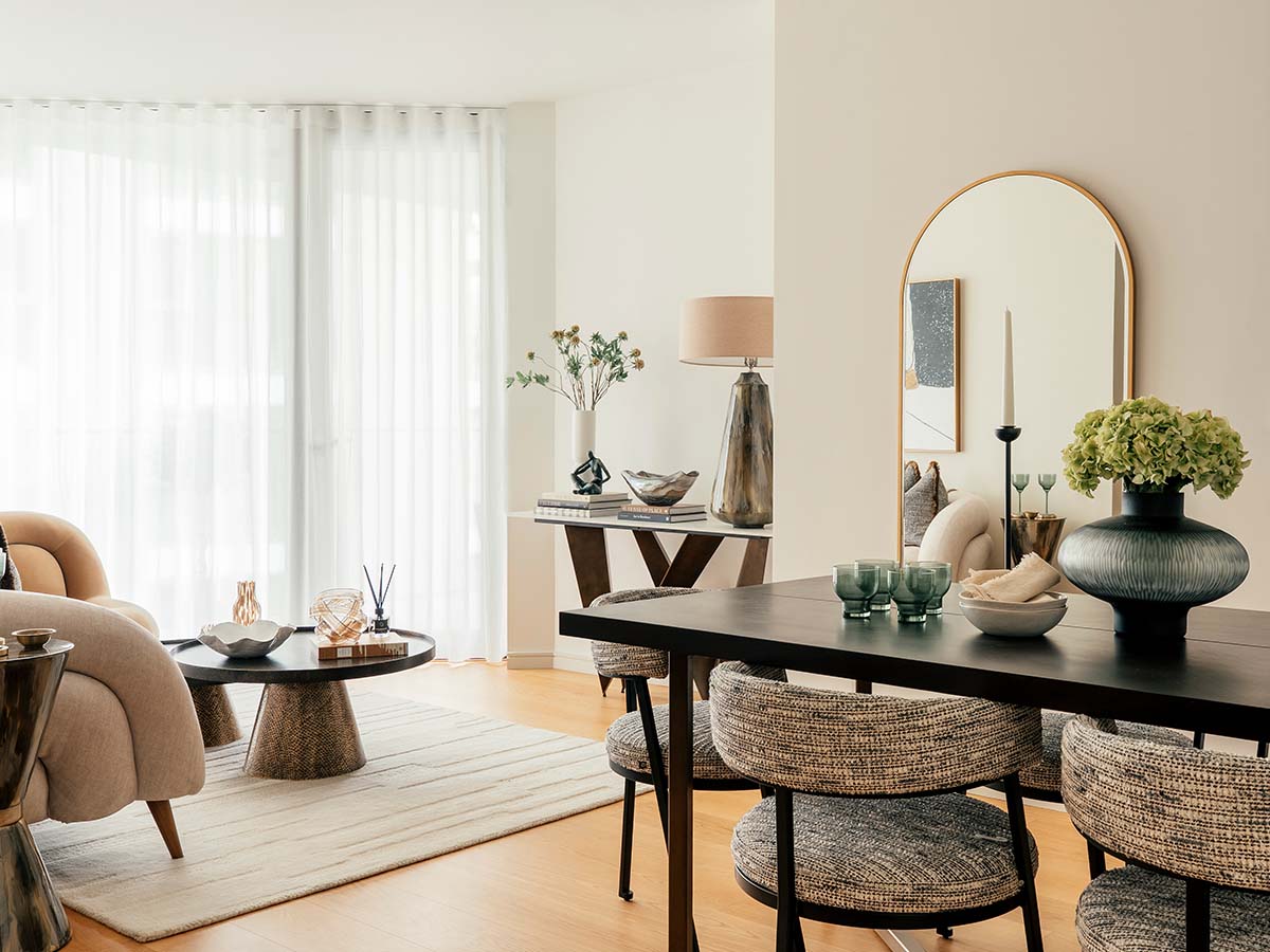 A contemporary dining area styled by InStyle Direct featuring a round dining table with neutral upholstered chairs, a curved grey sofa, and floor-to-ceiling windows offering panoramic city views.