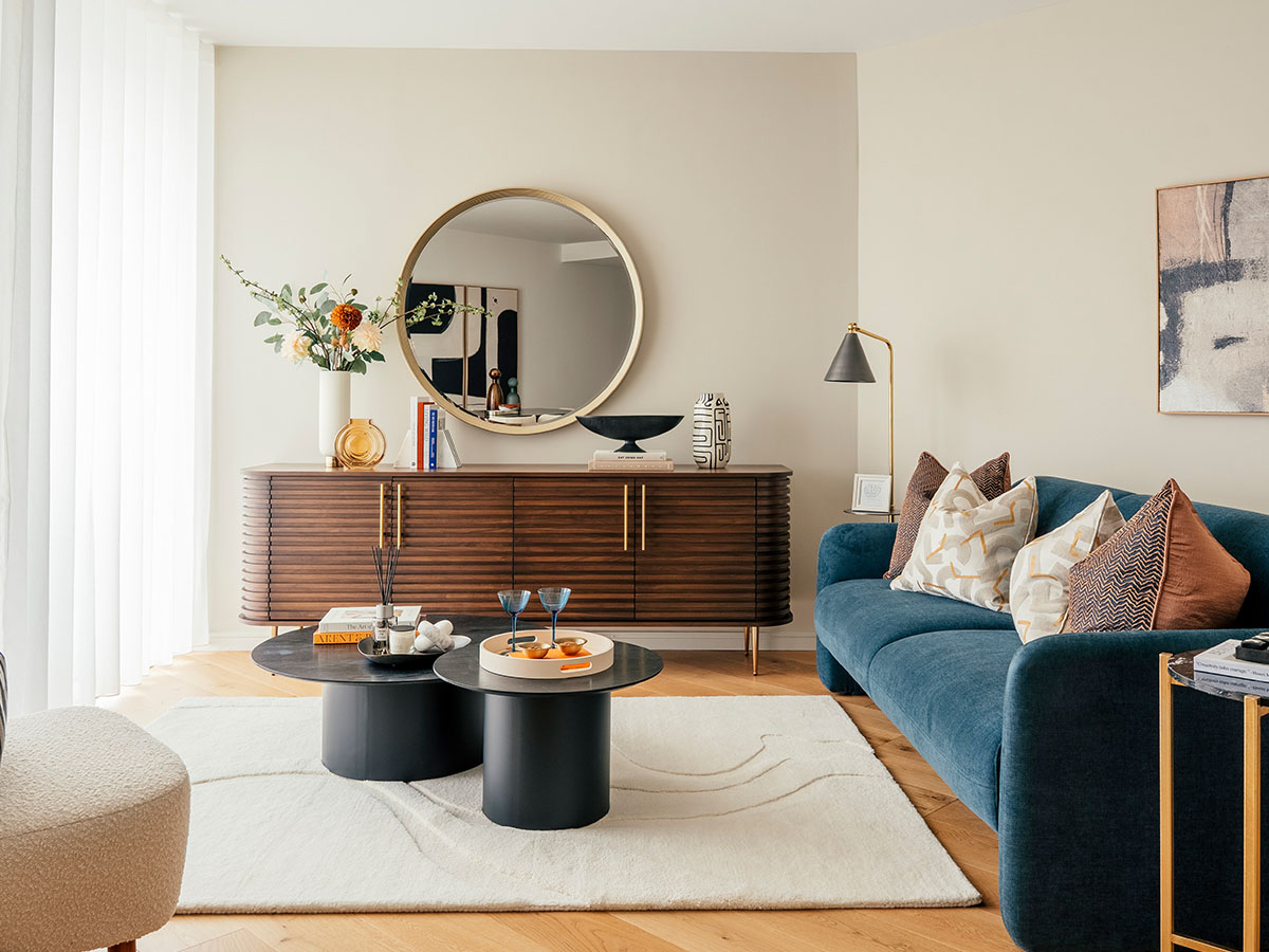 A luxury riverside apartment living area styled by InStyle Direct featuring a light grey sofa with velvet cushions, a black slat coffee table, and large indoor plants overlooking the Greenwich peninsula.
