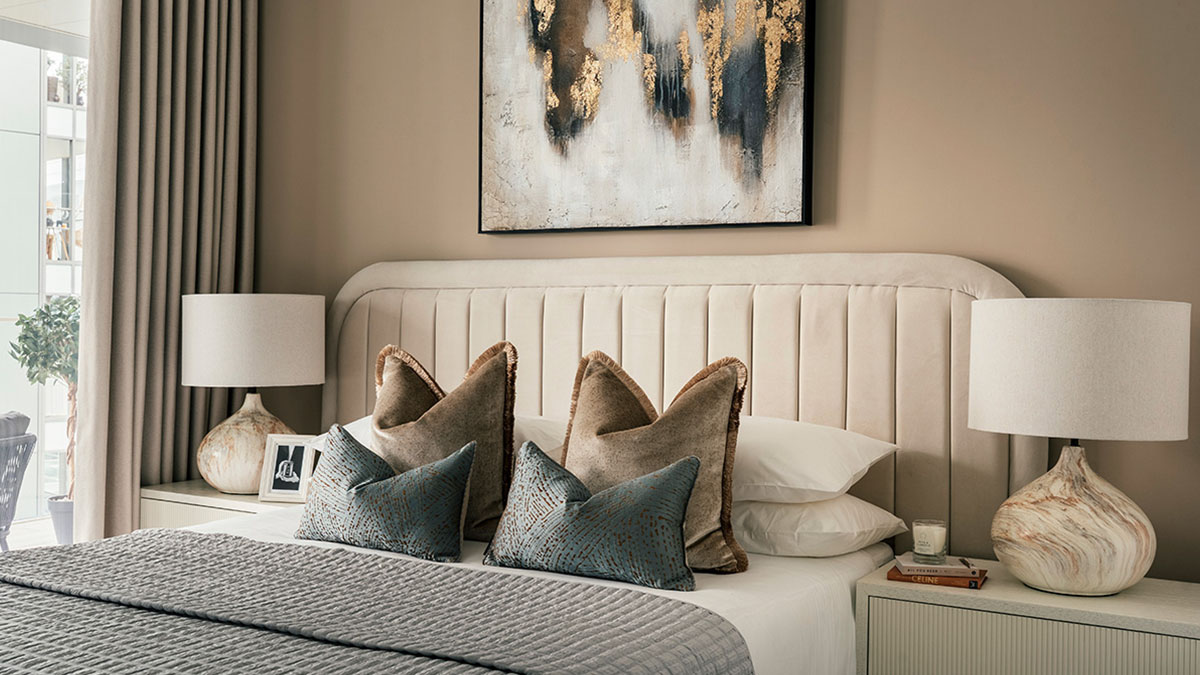 Modern bedroom with upholstered headboard, neutral tones, decorative cushions, bedside lamps, and abstract wall art