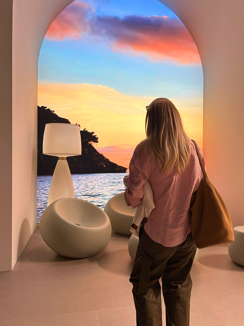 Woman standing near arched window overlooking sea at sunset with modern lounge chairs and floor lamp