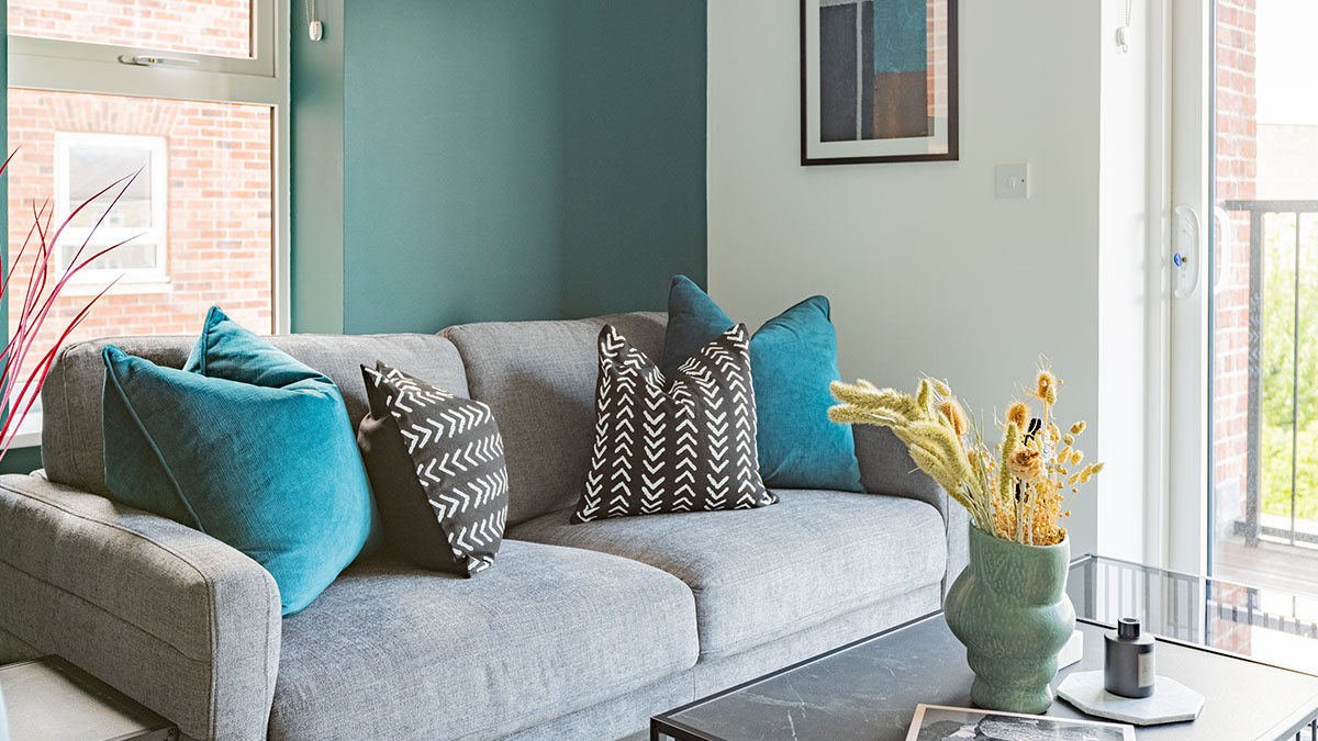 Modern living room with a grey sofa, teal accent cushions, patterned black-and-white pillows, and a decorative vase on a coffee table near large windows.