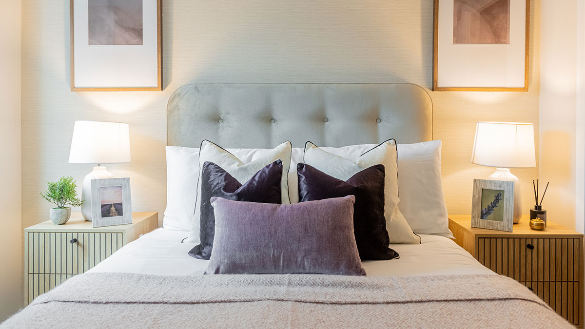 Stylish bedroom featuring a soft upholstered bed, purple accent cushions, bedside lamps and framed artwork.