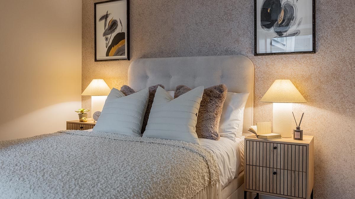 Elegant bedroom with upholstered headboard, layered cushions, bedside tables and warm table lamps.