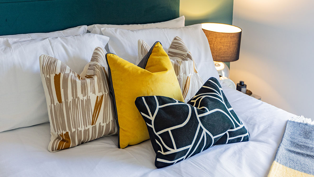 Stylishly dressed bed with layered patterned cushions in yellow, black, and neutral tones, complemented by a bedside lamp and upholstered headboard.