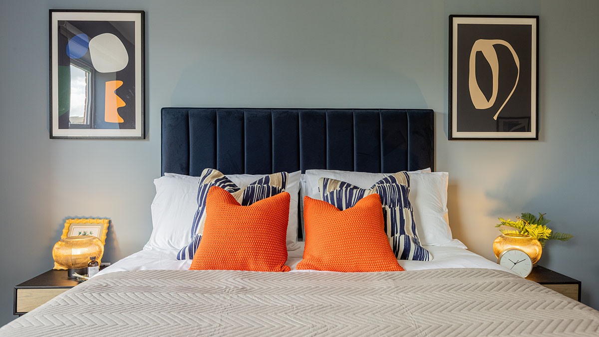 Contemporary bedroom featuring a navy upholstered headboard, vibrant orange accent cushions, abstract wall art, and illuminated bedside tables with decorative accessories.