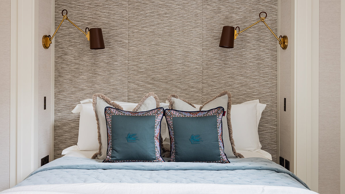 Luxury bedroom with an upholstered headboard, designer cushions, wall-mounted brass reading lights and soft blue bedding in a London apartment.
