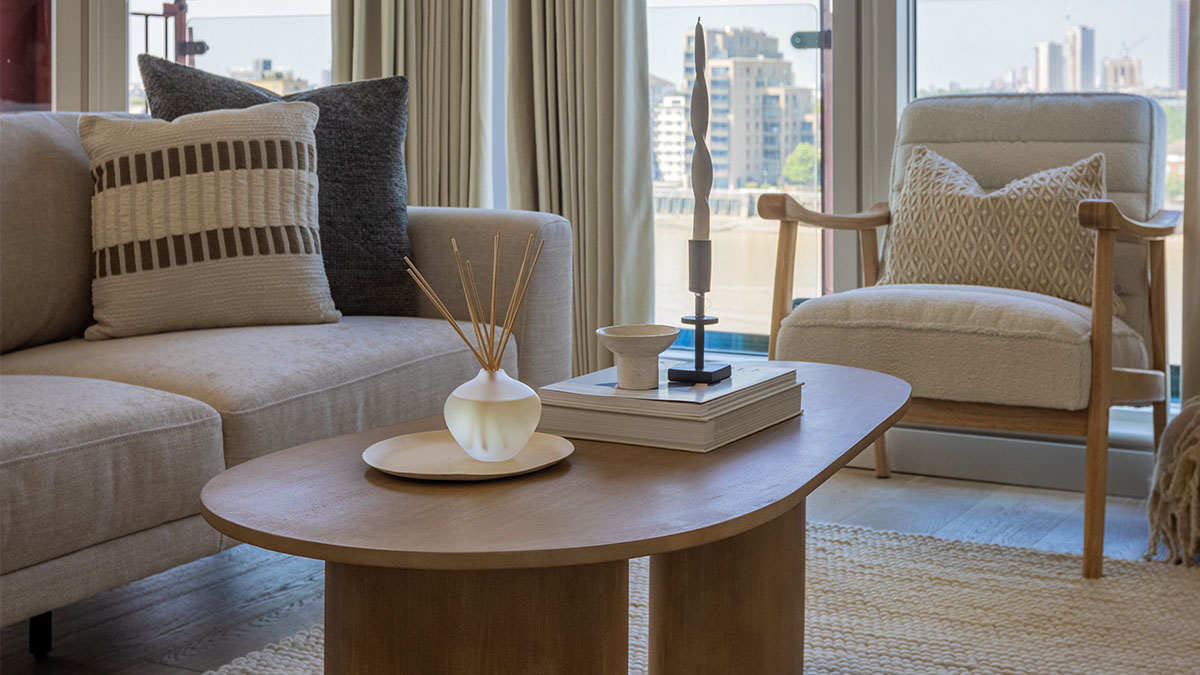 Close-up of a modern living room featuring a wooden coffee table, neutral sofa, accent chair, and floor-to-ceiling windows with riverside views.