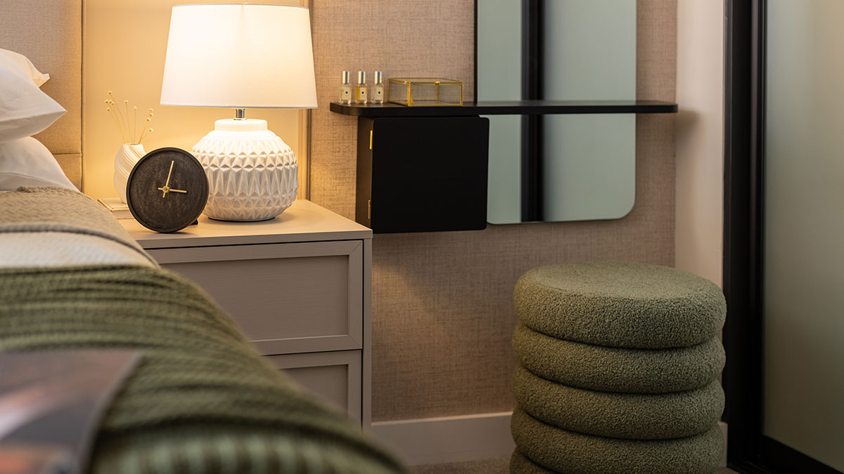 Bedroom dressing area with a wall-mounted vanity desk, mirror, bedside table and textured upholstered stool in a furnished apartment.