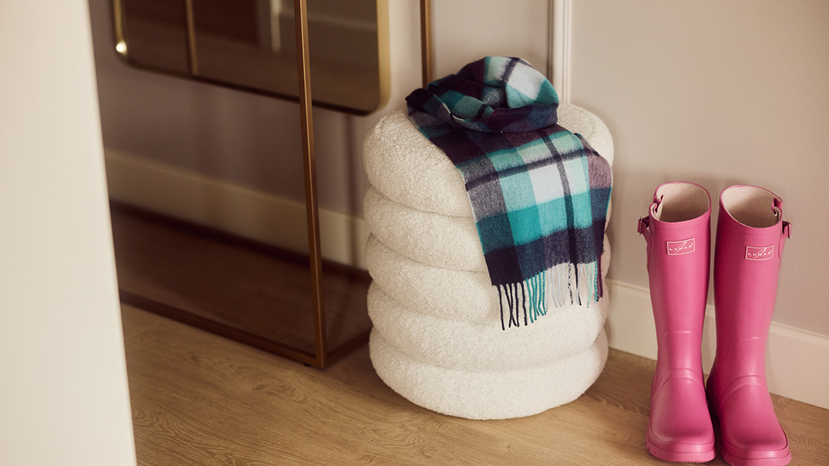 Stylish entryway featuring a boucle storage pouffe with a checked throw and pink wellington boots in a modern apartment.