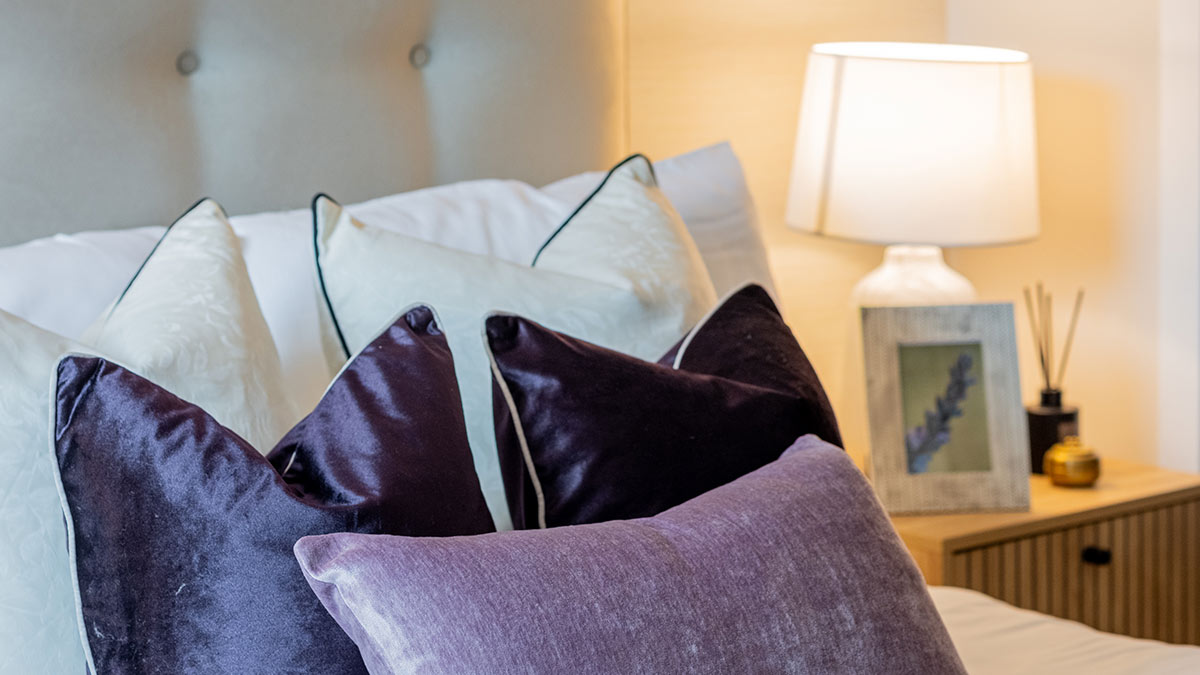 Luxury bedroom with white and purple decorative pillows, upholstered headboard, bedside lamp, and wooden nightstand