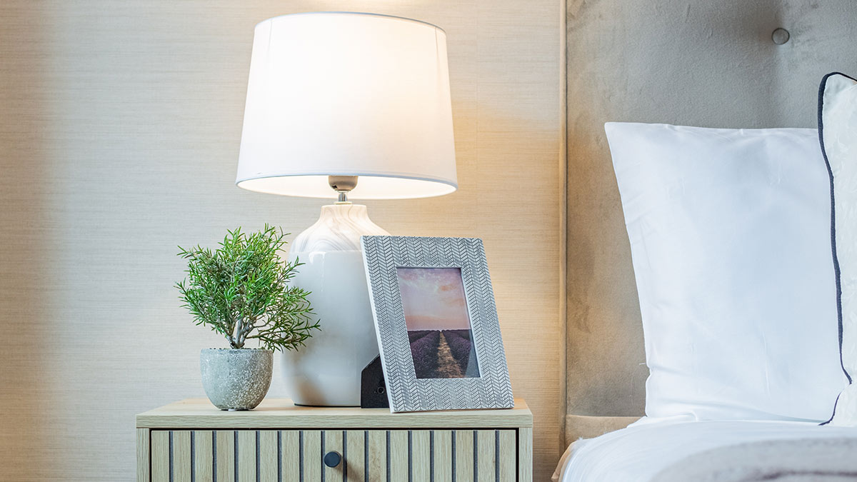 Cozy bedroom bedside table with a white lamp, potted green plant, framed artwork, and a neatly made bed.