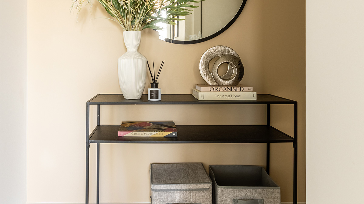 A stylish black console table against a warm beige wall, decorated with a white vase of greenery, reed diffuser, stacked books, and a metallic circular sculpture beneath a round mirror.