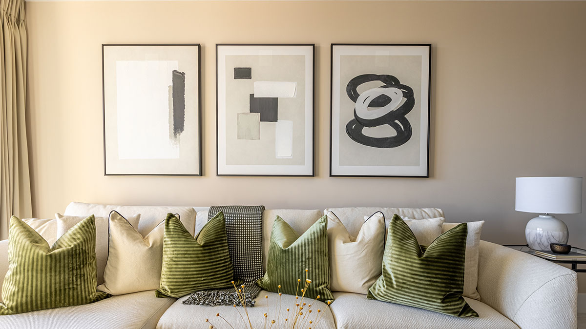Modern neutral living room with a cream sofa, green velvet cushions, abstract framed artwork, beige curtains, and a white table lamp.