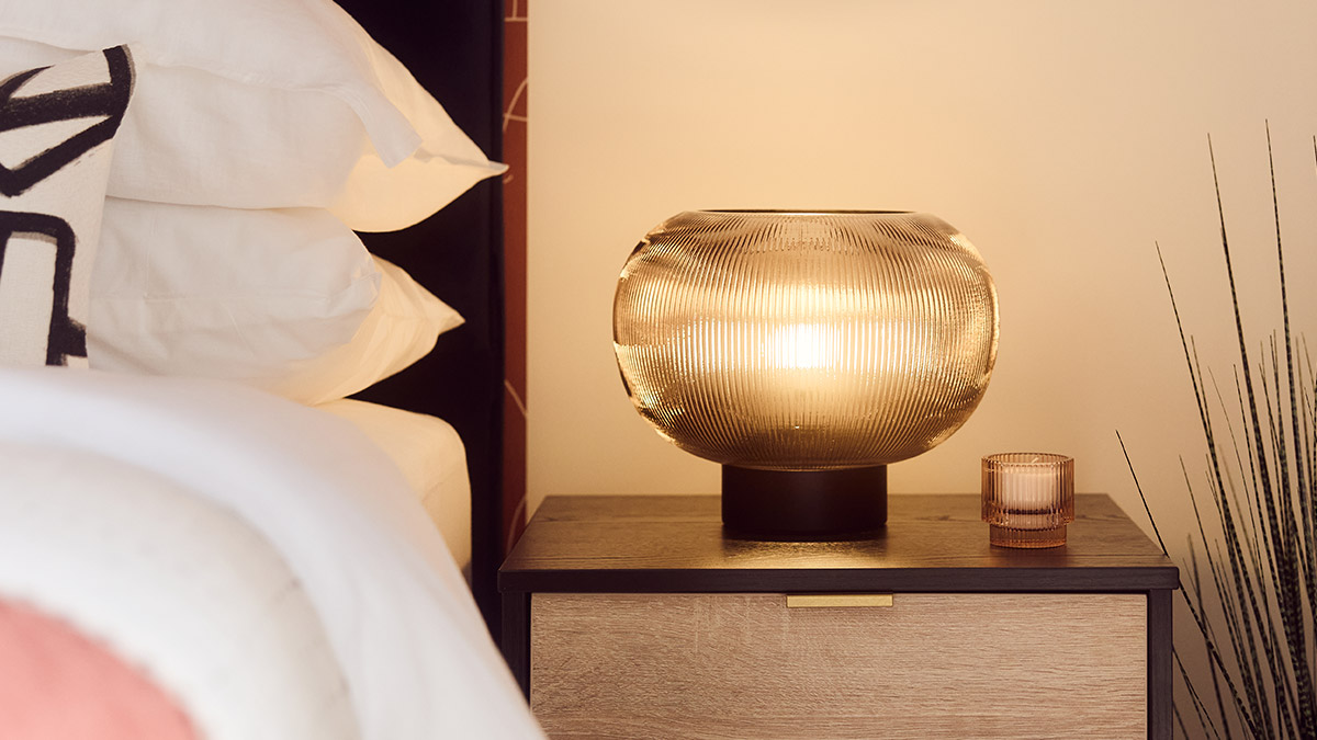 Warm bedside lighting with a ribbed glass table lamp, decorative candle and wooden bedside table beside a neatly styled bed