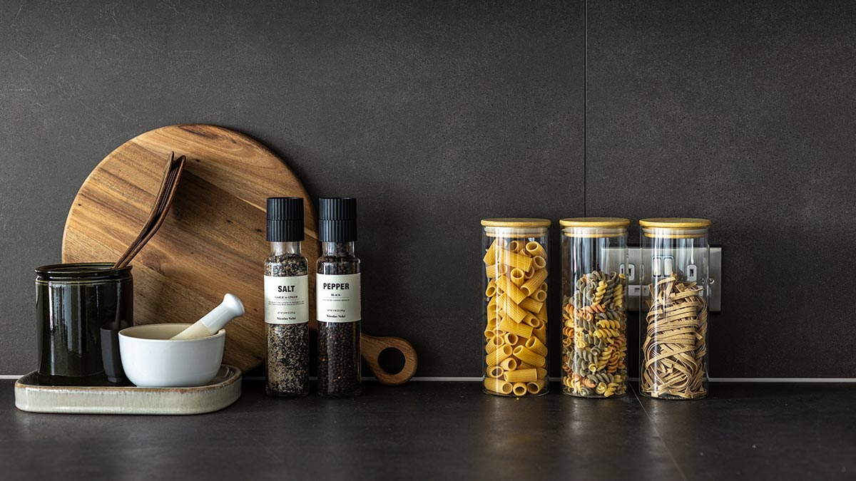 Dark stone kitchen countertop styled with wooden chopping boards, salt and pepper grinders, and glass jars of pasta.