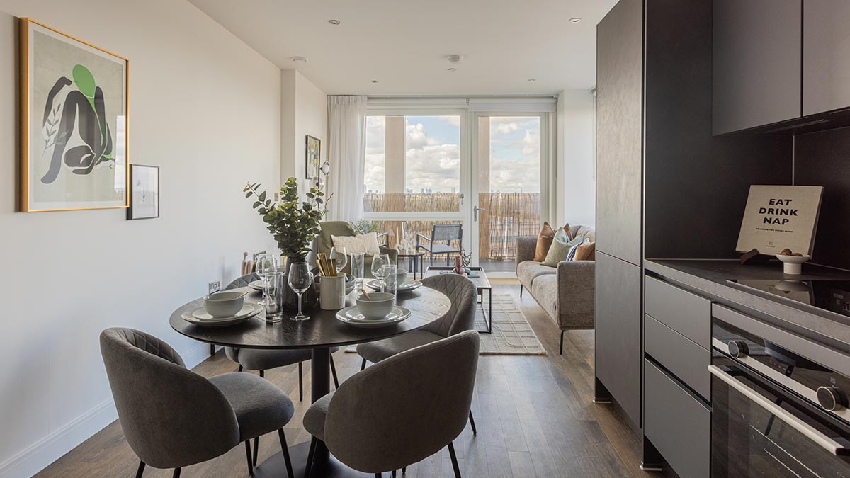 Modern apartment interior with dining area, grey upholstered chairs, dark kitchen cabinetry and balcony doors.